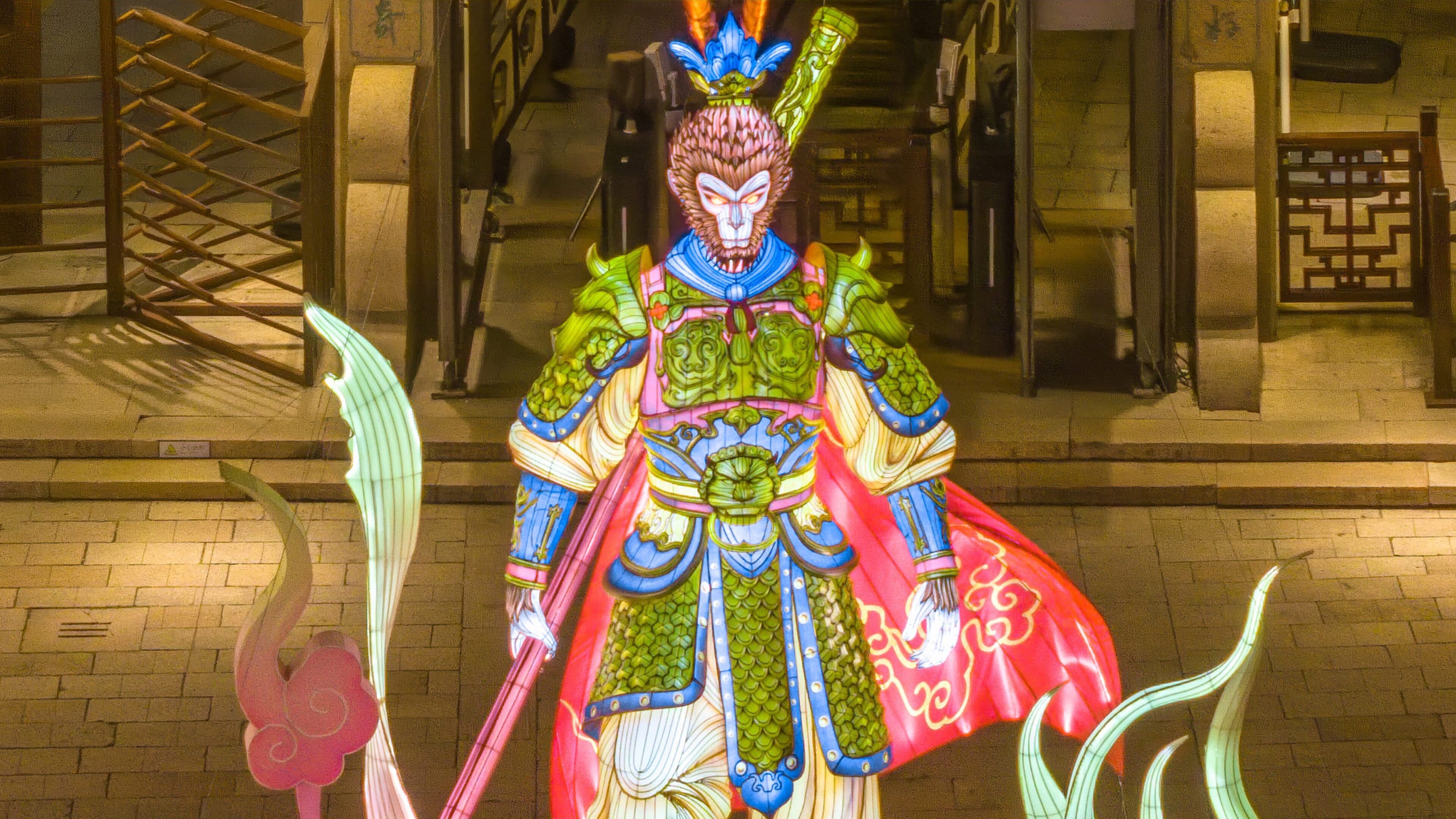 A large illuminated Sun Wukong lantern in armour holding a staff on a stone-paved street at night, wooden building behind
