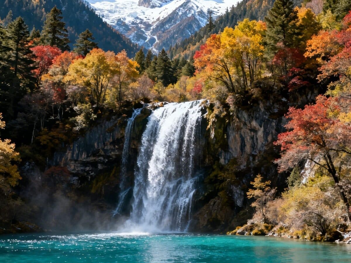 A waterfall dropping into a jade pool among autumn forest, with snow-dusted peaks behind the mist